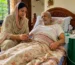 A woman checks the oxygen level of an elderly man lying in bed with an oxygen tube, in a home setting